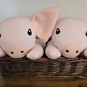 Puede incluir: Dos cerdos de peluche rosas con orejas grandes y ojos negros están en una cesta de mimbre marrón. Los cerdos tienen hocicos de color rosa claro y están hechos de material suave.