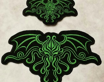 Op de afbeelding: Twee zwarte en groene geborduurde patches met een gestileerd Cthulhu-ontwerp. De grotere patch is ongeveer 25 cm breed en de kleinere patch is ongeveer 12,5 cm breed.