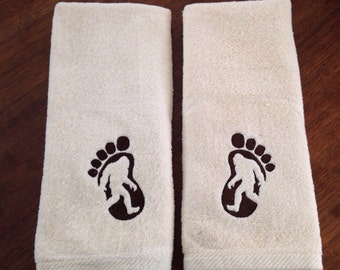 May include: Two white hand towels with brown embroidered footprints. Each footprint has a silhouette of a Bigfoot inside.