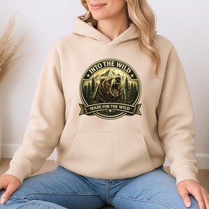 May include: A beige hoodie with a front pocket and a drawstring hood. The front features a circular graphic with a bear, mountains, and the text "INTO THE WILD" and "MADE FOR THE WILD" in a banner.