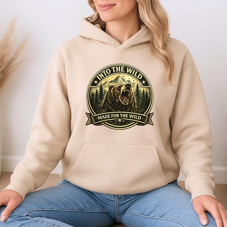 May include: A beige hoodie with a front pocket and a drawstring hood. The front features a circular graphic with a bear, mountains, and the text "INTO THE WILD" and "MADE FOR THE WILD" in a banner.