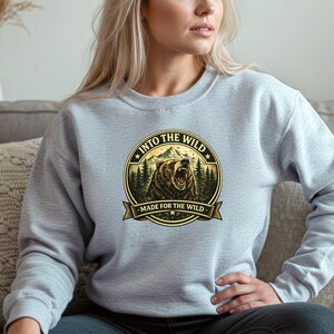 May include: A light gray sweatshirt featuring a circular graphic with a roaring bear, mountains, and pine trees. The text "INTO THE WILD" and "MADE FOR THE WILD" are displayed in a circular design.