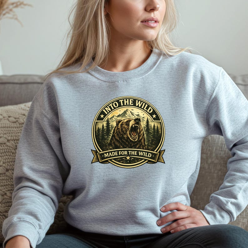 May include: A light gray sweatshirt featuring a circular graphic with a roaring bear, mountains, and pine trees. The text "INTO THE WILD" and "MADE FOR THE WILD" are displayed in a circular design.