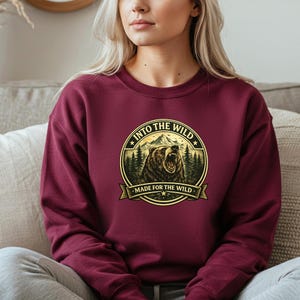 May include: Burgundy sweatshirt with a circular graphic featuring a roaring bear, mountains, and pine trees. Text reads "INTO THE WILD" and "MADE FOR THE WILD" in a gold color. The sweatshirt is a solid color.