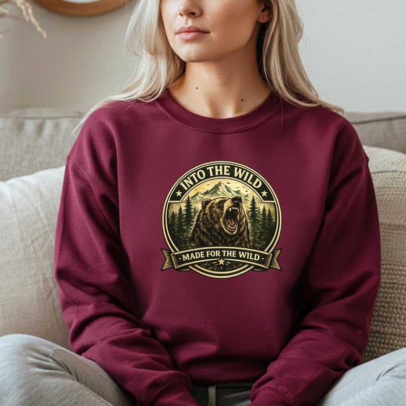 May include: Burgundy sweatshirt with a circular graphic featuring a roaring bear, mountains, and pine trees. Text reads "INTO THE WILD" and "MADE FOR THE WILD" in a gold color. The sweatshirt is a solid color.