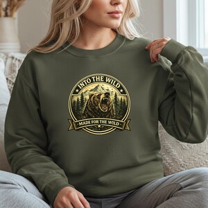 May include: Olive green sweatshirt with a circular graphic featuring a roaring bear, mountains, and trees. The text "INTO THE WILD" and "MADE FOR THE WILD" are displayed in a gold color. The sweatshirt is made of a soft material.
