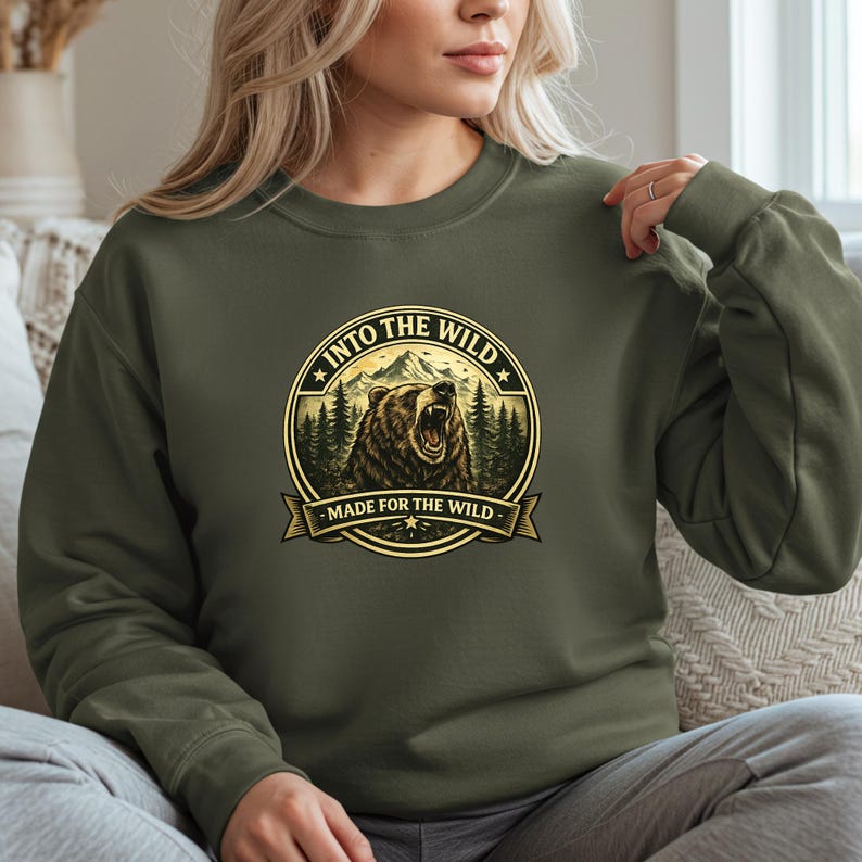 May include: Olive green sweatshirt with a circular graphic featuring a roaring bear, mountains, and trees. The text "INTO THE WILD" and "MADE FOR THE WILD" are displayed in a gold color. The sweatshirt is made of a soft material.