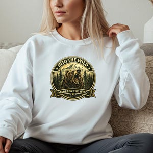 May include: White sweatshirt featuring a circular graphic with a bear, mountains, and trees. The text "INTO THE WILD" and "MADE FOR THE WILD" are displayed around the bear illustration. The sweatshirt is a casual style.