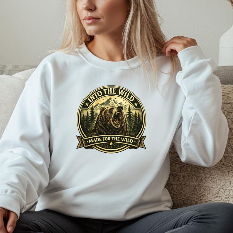 May include: White sweatshirt featuring a circular graphic with a bear, mountains, and trees. The text "INTO THE WILD" and "MADE FOR THE WILD" are displayed around the bear illustration. The sweatshirt is a casual style.