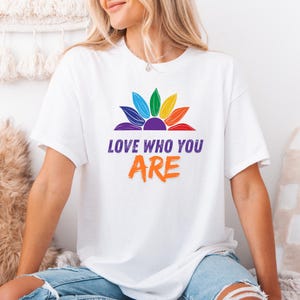 Può includere: T-shirt bianco con un motivo a girasole arcobaleno. Il testo "LOVE WHO YOU ARE" è stampato sotto in viola e arancione. Un capo di abbigliamento casual.