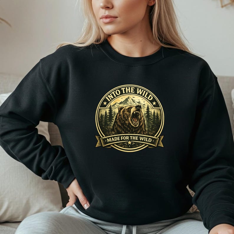 May include: Black sweatshirt featuring a circular graphic with a bear, mountains, and trees. The graphic includes the text "INTO THE WILD" and "MADE FOR THE WILD" in a gold color. The sweatshirt is a solid black color.