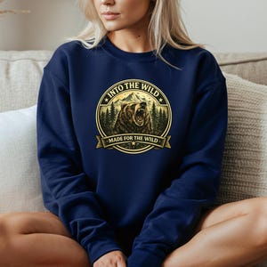 May include: Navy blue sweatshirt featuring a circular graphic with a bear, mountains, and trees. The text reads "INTO THE WILD" and "MADE FOR THE WILD" in a gold color. The sweatshirt is a solid navy blue color.