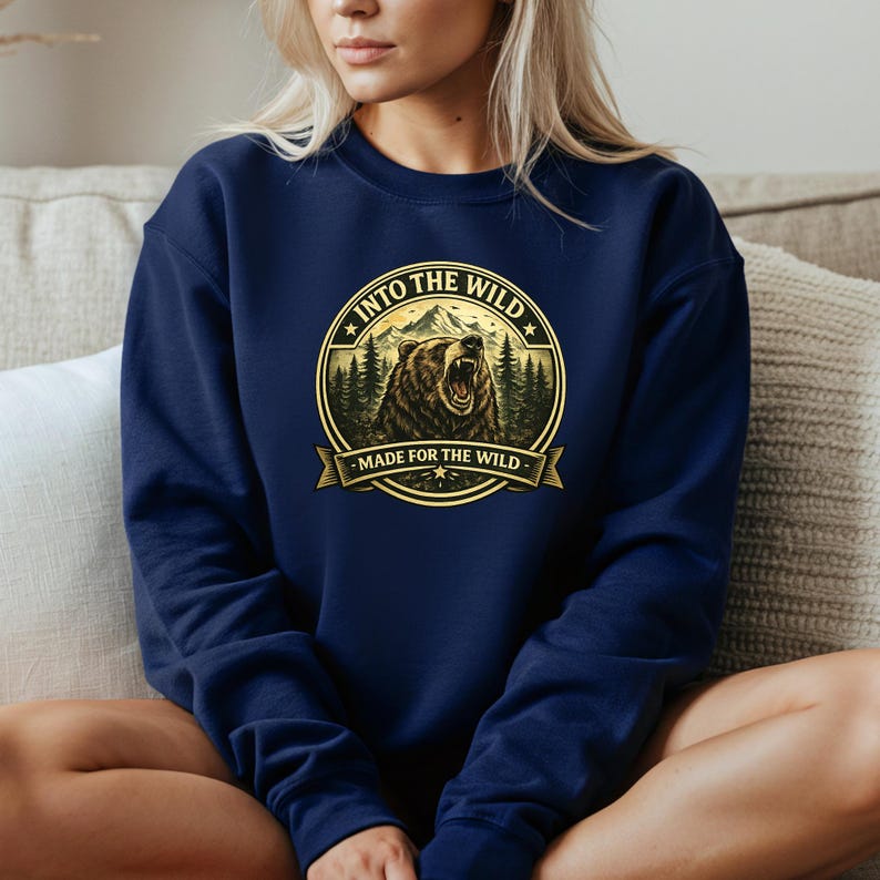 May include: Navy blue sweatshirt featuring a circular graphic with a bear, mountains, and trees. The text reads "INTO THE WILD" and "MADE FOR THE WILD" in a gold color. The sweatshirt is a solid navy blue color.
