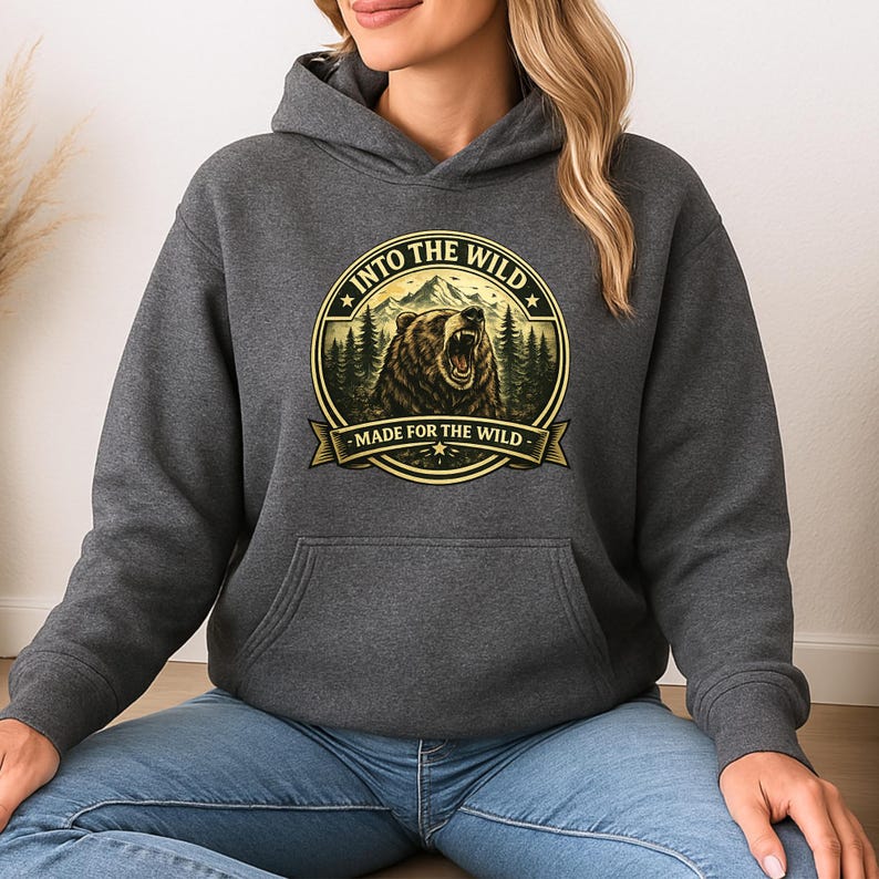 May include: A charcoal gray hoodie with a front pocket and a drawstring hood. The front features a circular graphic with a roaring bear, mountains, and the text "INTO THE WILD" and "MADE FOR THE WILD" in a gold color.