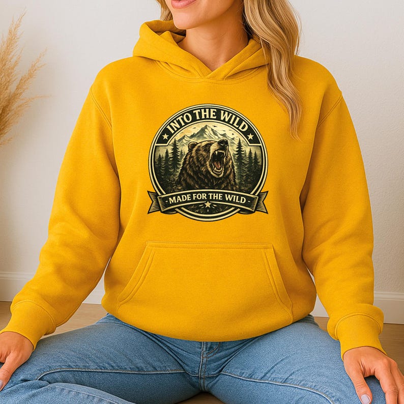 May include: Mustard yellow hoodie with a front pocket and a drawstring hood. The graphic on the front features a bear roaring in front of a mountain and forest scene, with the text "INTO THE WILD" and "MADE FOR THE WILD".