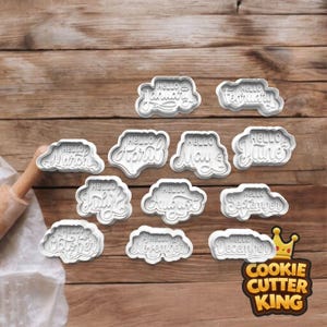 May include: A set of white plastic cookie cutters, each featuring the word "Hello" and a month of the year. The cutters are displayed on a wooden surface. A rolling pin is visible on the left.