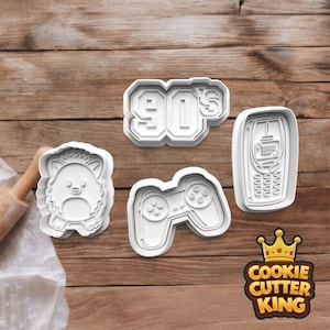 May include: Four white plastic cookie cutters: a retro mobile phone, a games controller, a cartoon animal, and the number "90's". The cutters are on a wooden surface.
