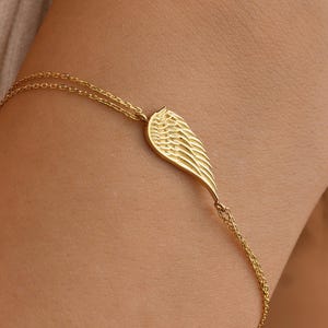 Pode incluir: Uma delicada pulseira dourada com um berloque de asa. A asa &eacute; detalhada com linhas finas, e a pulseira tem uma corrente fina. A joia &eacute; usada no pulso, mostrando o design elegante. A pulseira &eacute; um acess&oacute;rio bonito.