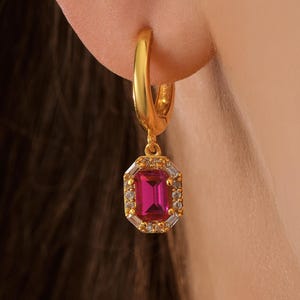 May include: Gold hoop earring with a rectangular, bright pink gemstone pendant. The pendant is framed by a halo of small, clear stones. The earring is shown on a person's ear.