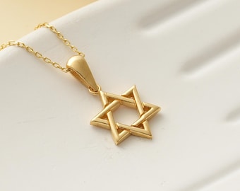 May include: A gold-colored Star of David pendant necklace. The pendant is a six-pointed star, and the necklace is displayed on a white hand-shaped ceramic dish. The background includes yellow flowers.
