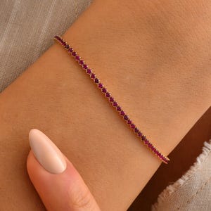 May include: A delicate gold-tone tennis bracelet featuring a continuous row of small, vibrant magenta-pink circular gemstones set in a thin, polished metal chain that encircles the wrist.