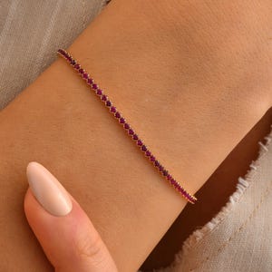 Könnte beinhalten: Ein zartes goldenes Armband, besetzt mit kleinen, leuchtend roten Edelsteinen. Das Armband ist dünn und elegant und weist eine durchgehende Reihe roter Steine auf. Der Schmuck wird an einem Arm präsentiert, um sein Design und seine Farbe hervorzuheben.