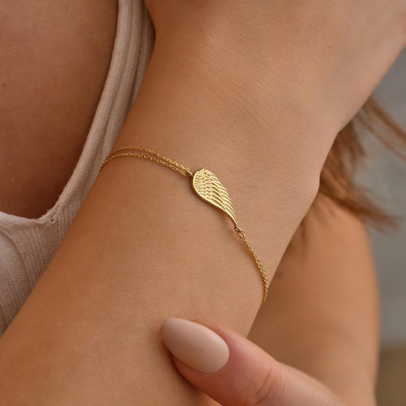 Pode incluir: Uma delicada pulseira dourada com um berloque de asa. A asa &eacute; detalhada com linhas finas, e a pulseira tem uma corrente fina. A joia &eacute; usada no pulso, mostrando o design elegante. A pulseira &eacute; um acess&oacute;rio bonito.