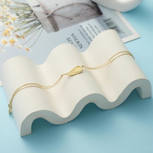 Pode incluir: Um delicado colar dourado com um pingente de asa. O colar &eacute; exibido em uma superf&iacute;cie branca ondulada sobre um fundo azul claro. O pingente de asa &eacute; detalhado e a corrente &eacute; fina.