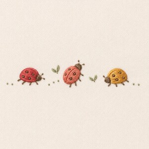 May include: An embroidery design on a light fabric featuring three ladybirds in a horizontal row. The ladybirds are coloured red, coral, and yellow, each with dark brown spots and antennae. Small green leaf accents and tiny dots are interspersed.