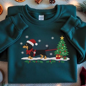 May include: A dark teal sweatshirt featuring a Christmas-themed dachshund design. The dog wears a Santa hat and a red bow, pulling a decorated Christmas tree on a leash. Snowflakes and festive greenery complete the holiday scene.