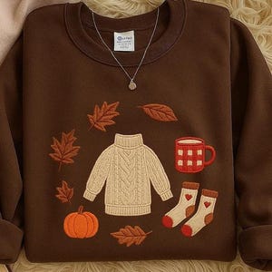 May include: A brown sweatshirt with embroidered autumn-themed designs. The design includes a cream-colored cable-knit sweater, a pumpkin, fall leaves, a red mug, and socks. A silver necklace with a small pendant is also visible.