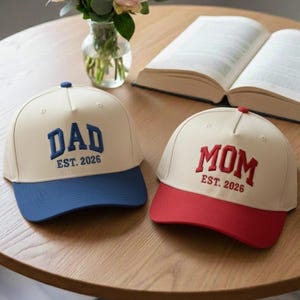 May include: Two baseball caps with the words "DAD" and "MOM" embroidered on them. The "DAD" cap has a blue brim and lettering, while the "MOM" cap has a red brim and lettering. Both caps have "EST. 2026" embroidered below the main text.