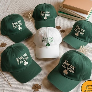 May include: Several baseball caps in shades of green and one in white, all featuring the embroidered text "Kiss me I'm Irish" and a shamrock. The caps are displayed on a textured surface, with shamrock decorations.