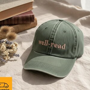 May include: A khaki green baseball cap featuring the embroidered words "well-read" in pink. The cap has a curved peak and a classic six-panel design. The background includes books and dried flowers.