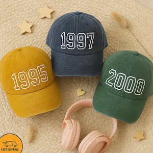 May include: Three baseball caps in mustard yellow, navy blue, and forest green, each embroidered with a different year in white. A pair of pink headphones and star-shaped decorations are also visible.