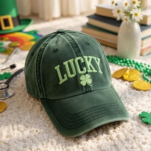 May include: A green baseball cap with the word "LUCKY" embroidered in light green, along with a shamrock. The cap is on a white carpet, with gold coins and a green bead necklace nearby. A green leprechaun hat is in the background.
