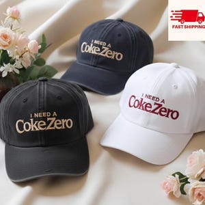 Puede incluir: Tres gorras de béisbol en azul marino, negro y blanco, cada una bordada con "I NEED A Coke Zero". Las gorras están dispuestas sobre una superficie clara con acentos florales. La gorra azul marino tiene bordados blancos, la negra bordados beige y la blanca bordados rojos.