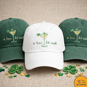 May include: Three baseball caps are shown: two green and one white. Each cap has an embroidered martini glass with olives and a shamrock, and the text "a tini bit irish". Shamrocks and gold coins are scattered around the hats as decoration.
