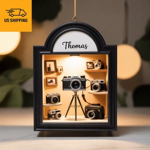 May include: A black framed display case featuring miniature cameras and framed photos. The name "Thomas" is written in cursive at the top. A central camera sits on a small tripod. The display is lit from within.