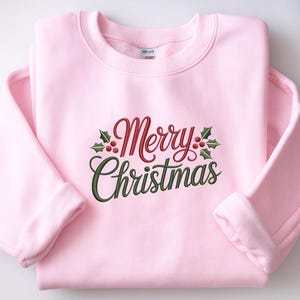 May include: A light pink sweatshirt with the embroidered text "Merry Christmas" in red and green script, accented with holly leaves and berries. The sweatshirt has a crew neck and long sleeves.
