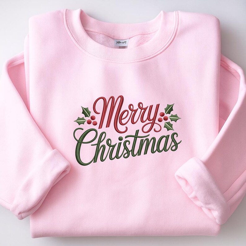 May include: A light pink sweatshirt with the embroidered text "Merry Christmas" in red and green script, accented with holly leaves and berries. The sweatshirt has a crew neck and long sleeves.