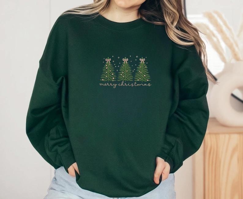 May include: Dark green sweatshirt with embroidered Christmas trees and the text "merry christmas" in a delicate script. The trees are decorated with tiny lights and bows, creating a festive holiday design.