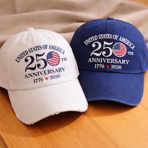 May include: Two baseball caps, one white and one navy blue, are displayed. Both caps feature the text "UNITED STATES OF AMERICA 250TH ANNIVERSARY 1776-2026" with an American flag design. The white cap has a distressed brim.