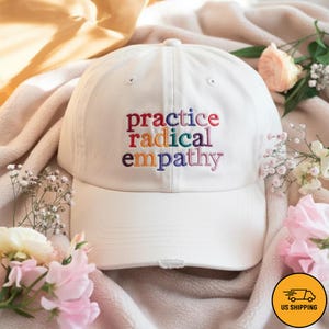 Puede incluir: Gorra de béisbol blanca con el texto bordado "practice radical empathy" en letras coloridas. El sombrero está hecho de un material suave y tiene una visera curva. Un accesorio casual.
