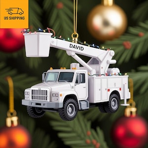 Op de afbeelding: Witte kerstversiering in de vorm van een utility truck met de naam "DAVID" op de giek. De truck heeft een hoogwerker en is versierd met kleurrijke lichtjes. De versiering hangt aan een gouden draad.