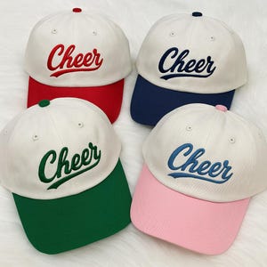 Op de afbeelding: Vier baseballpetten met het woord "Cheer" geborduurd op de voorkant. Elke pet heeft een witte kroon en een gekleurde klep. Kleuren zijn rood, marineblauw, groen en roze. De petten zijn een sportief accessoire.