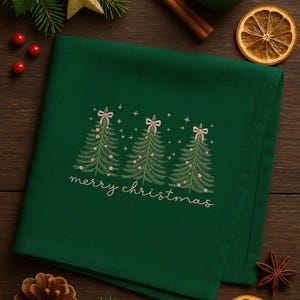 May include: A dark green Christmas-themed cloth napkin with embroidered Christmas trees, pink bows, and the words "merry christmas". The napkin is surrounded by holiday decorations, including a gold star and pine cones.