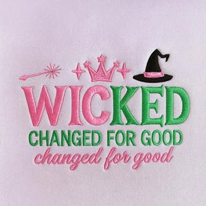Puede incluir: Diseño bordado con la palabra "WICKED" en rosa y verde, y "CHANGED FOR GOOD" en verde y rosa. Incluye una corona, estrellas, un sombrero de bruja y una varita sobre un fondo rosa claro.