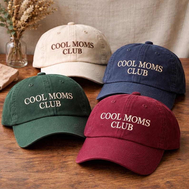 May include: Four baseball caps in cream, navy, green, and burgundy. Each cap has the embroidered text "COOL MOMS CLUB" in a contrasting color. The caps are arranged on a wooden surface.