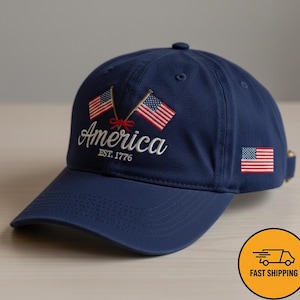May include: A navy blue baseball cap with the word "America" embroidered in white, along with two American flags and the text "EST. 1776". The cap also features a small American flag on the side.